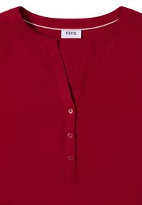 Rød skjorte med knaplukning, der har V-hals, korte ærmer og fem matchende knapper langs stolpen. Glat stof med et enkelt design.