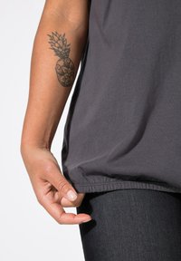Top grigio a maniche corte realizzato in tessuto morbido, presenta una vestibilità ampia e un orlo arricciato. Una mano tiene l'orlo vicino a un paio di jeans in denim scuro.