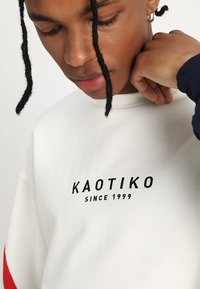 Hvit sweatshirt med svart brodert tekst "KAOTIKO SINCE 1999." Har en ribbestrikket krage og kontrasterende rød stripe på ermene.