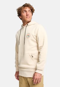 Sudadera beige con bolsillo frontal, capucha ajustable con cordón y parche de logo que presenta un sol y olas. Tejido suave y texturizado.