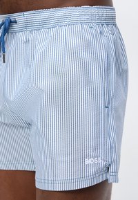 Άνδρας που φοράει σορτς με στενές κάθετες ρίγες σε ανοιχτό μπλε και λευκό, με ελαστική μέση και μπλε κορδόνι, με το λογότυπο "BOSS" στο αριστερό πόδι.