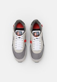 Gray sneakers with a beige upper, white laces, red accents, and a graphic logo on the tongue. Textured materials and a lightweight design.