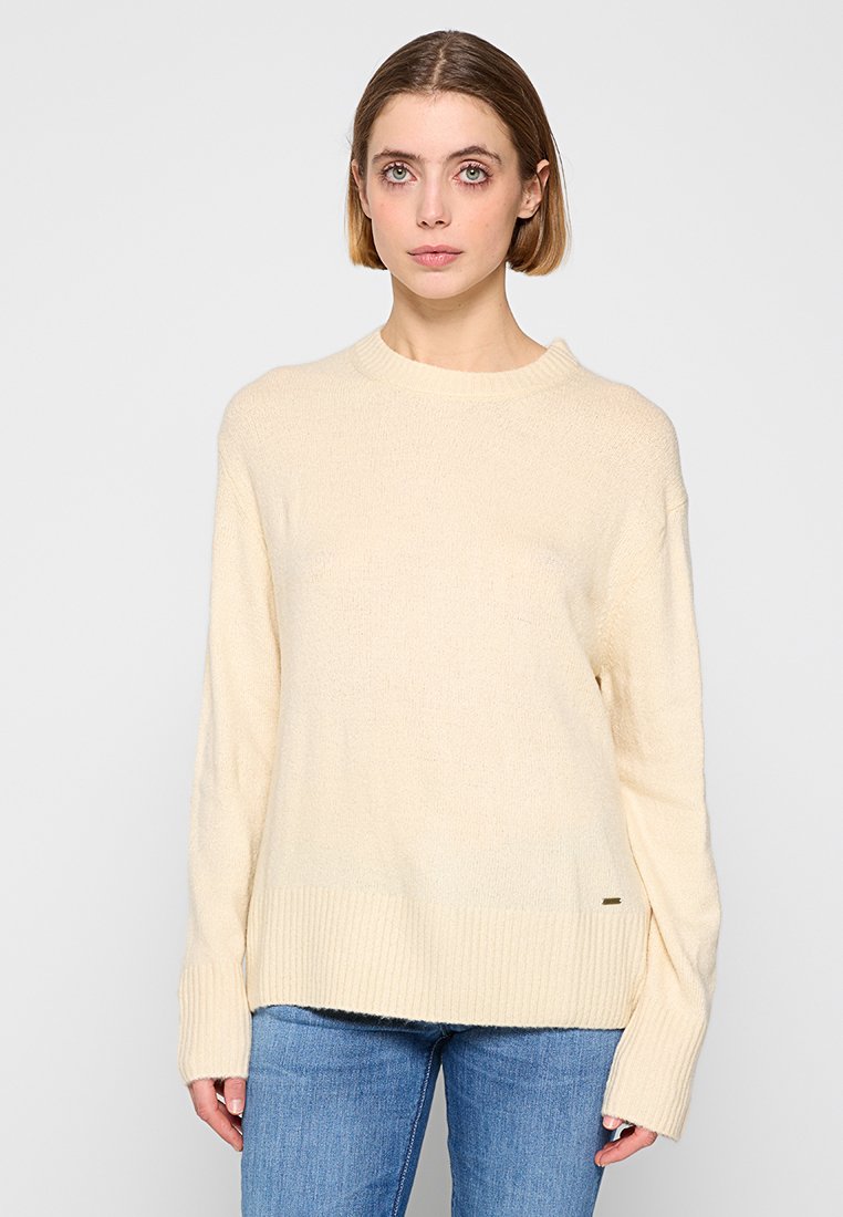 Pull en tricot beige avec un col rond et un ourlet côtelé. Manches longues et texture douce, associé à un jean bleu. Détail logo subtil à l'ourlet.