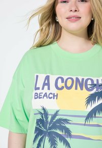 Camiseta de gran tamaño en verde claro con un estampado gráfico de palmeras, un sol y el texto "LA CONCHA BEACH" en colores contrastantes y en negrita.