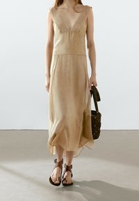 Nainen, jolla on yllään beige hihaton midi-mekko syvällä V-kaula-aukolla, ruskeat hihnasaappaat, kantaa ruskeaa käsilaukkua ja kävelee sileällä lattialla.