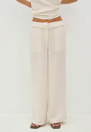 Vrouw die crèmekleurige, hooggetailleerde, wijde broek met trekkoord en zwarte sandalen draagt, staand tegen een effen witte achtergrond.