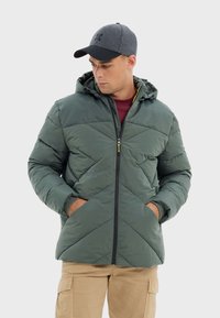 Veste isolante verte avec un motif de matelassage diagonal, capuche et fermeture éclair à l'avant. Elle est dotée de poignets élastiques et de poches latérales.