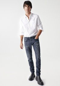 Camicia bianca con bottoni, jeans skinny blu scuro e stivaletti neri alla caviglia. Tessuto liscio, design aderente e motivi semplici in tutta la parte.