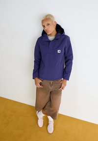 Veste à capuchon violette avec une poche avant, t-shirt gris en dessous, shorts large marron, et baskets roses avec des chaussettes blanches.