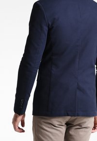 Marinblazer i textil med skräddarsydd passform, med tvåknäppt stängning och tre knappar på varje ärm; matchad med ljusbeige byxor.