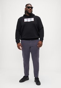 Black hoodie with white logo, gray pants, and black sneakers; model wears dark sunglasses; smooth fabric, relaxed fit, ample pockets.