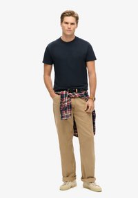 Camiseta azul marino, chinos beige, camisa de franela a cuadros atada a la cintura, zapatillas beige y un cinturón negro, con un reloj de pulsera en la muñeca izquierda.