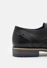 Schwarzer Lederschuh mit glatter Textur, ausgestattet mit einer schichtweisen Sohle in Tan und Grau sowie einem kleinen farbigen Akzent an der Seite.