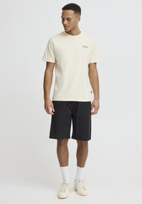 Kremhvid bomulds-t-shirt med lille logo, kombineret med sorte shorts med to forlommer. Hvide sokker og lyse sneakers fuldender outfittet.