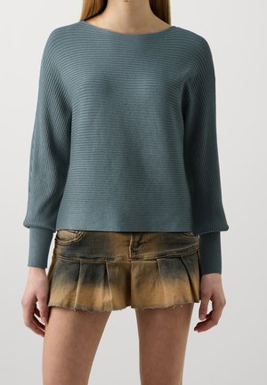 Femme portant un pull côtelé turquoise à manches longues et une mini-jupe plissée marron avec un motif délavé, debout devant un fond uni.