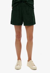 Persona vestida con pantalones cortos texturizados de color verde oscuro y zapatillas de color crema, de pie sobre un fondo blanco.