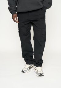 Pantalones cargo negros de mezcla de algodón; presentan bolsillos laterales, ajuste holgado y una textura suave. Combinados con zapatillas deportivas en gris y blanco.