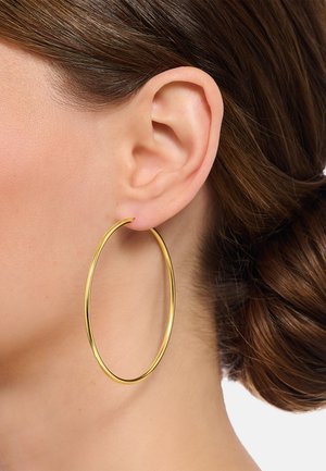 Nahaufnahme eines Frauenohrs, geschmückt mit einem großen, dünnen goldenen Creolenohrring und braunem nach hinten gestyltem Haar.