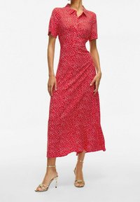 Robe rouge avec un design boutonné, manches courtes, pois blancs et jupe fluide. Associée à des talons dorés à brides.