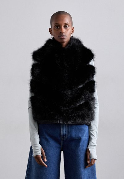 Gilet en fausse fourrure noire avec un design en couches, présentant une texture brillante. Associé à un haut à manches longues gris clair et un jean bleu à jambes larges.