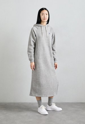 Grå hoodieklänning med långa ärmar, ribbade muddar och dragsko i huvan. Har en sidslits och är matchad med vita sneakers och grå strumpor.