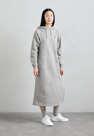 Grijze hoodie jurk met lange mouwen, ribgebreide manchetten en een trekkoordkap. Heeft een zijopening en is gecombineerd met witte sneakers en grijze sokken.