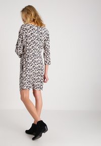 Lyhythihainen mekko vaaleassa kermassa, mustilla lehtikuvioilla, vyötäröltä istuva, polven ylle ulottuva pituus, takana avainreiän yksityiskohta.