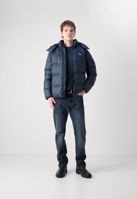 Marine pufferjack met een hoge kraag, gedragen over een donkere trui, gecombineerd met blauwe jeans en zwarte laarzen, met geborduurde logo-details.