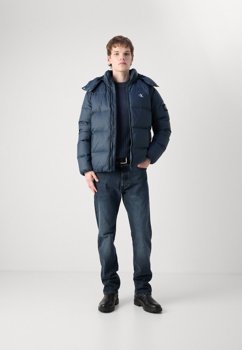 Marineblaue Steppjacke mit hohem Kragen, getragen über einem dunklen Pullover, kombiniert mit blauen Jeans und schwarzen Stiefeln, mit gestickten Logodetails.