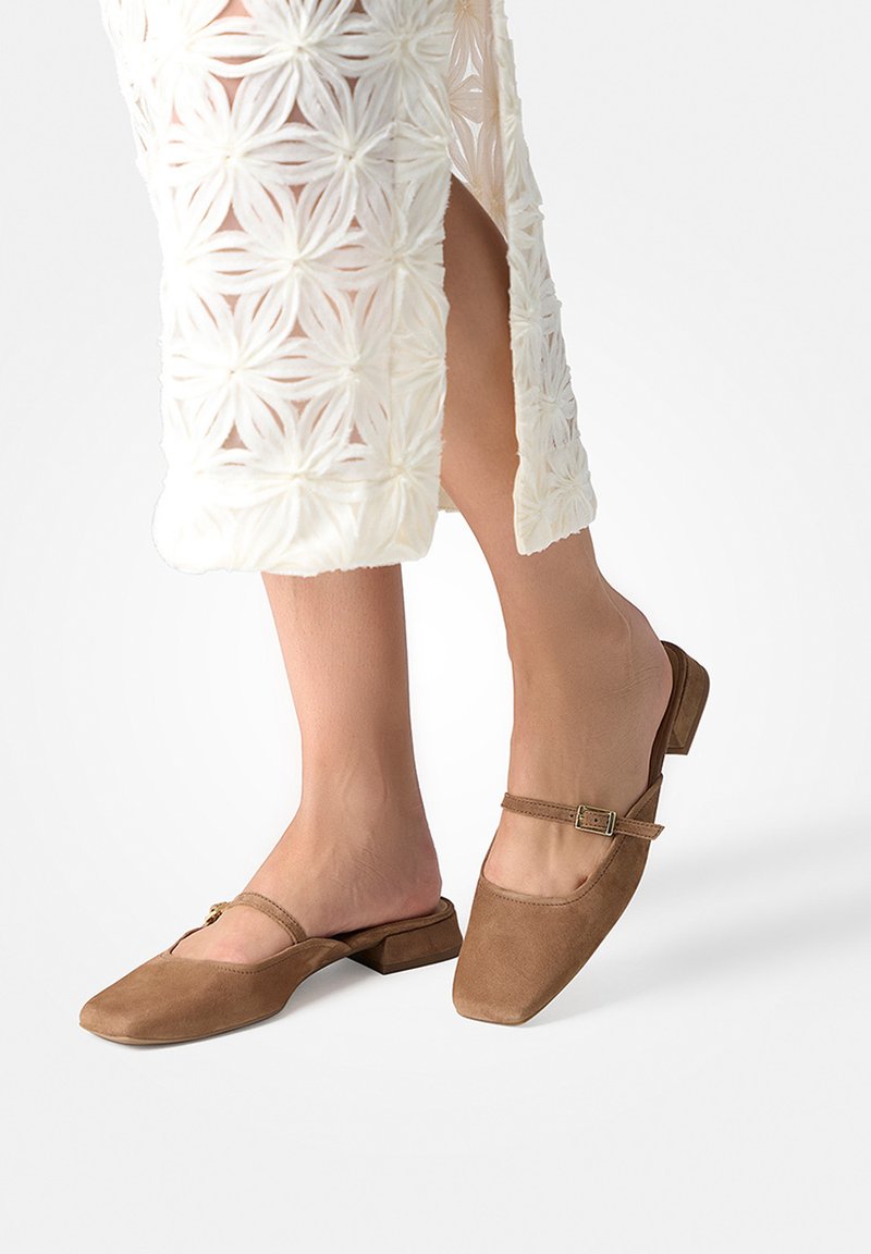 Füße in braunen Wildleder-Schlupfschuhen mit quadratischer Spitze, dünnem Riemen und Schnalle, kombiniert mit weißem floralen Spitzenrock mit Seitenschlitz.