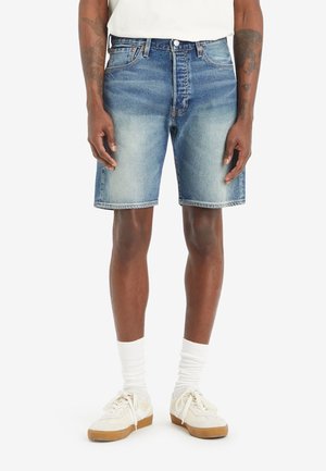 Homme portant un short en jean bleu délavé, des chaussettes blanches et des baskets beiges, debout, les bras détendus le long du corps, sur fond blanc.