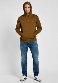 Felpa marrone con cappuccio, dotata di tasca frontale e cordoncino regolabile. Indossata con jeans di denim blu e sneakers bianche con accenti grigi.