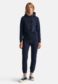 Marineblå hettegenser og bukse med teksturert logo på genseren, elastiske mansjetter og snor i midjen, sammen med lyse sneakers.