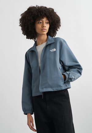 Mladá žena s kučeravými vlasmi, oblečená v modrej vetrovke North Face so zapínaním na zips, svetlosivej košeli a čiernych nohaviciach, stojaca na neutrálnom pozadí.