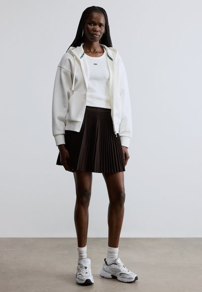 Sweat à capuche blanc à fermeture éclair, haut blanc ajusté, jupe plissée marron, baskets blanches et noires, chaussettes à la cheville ; tissu lisse et lignes de design nettes.
