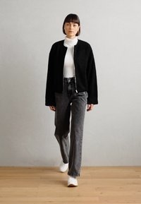 Giubbotto bomber nero, strutturato, con zip sopra un dolcevita bianco. Indossato con pantaloni grigi a vita alta e sneakers bianche. Design semplice e moderno.