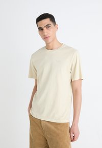 Crème-kleurige katoenen t-shirt met een ronde hals en korte mouwen. Heeft een klein logo in lichtblauw op de borst. Eenvoudige, relaxte pasvorm.