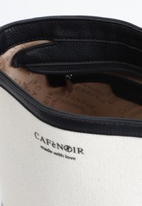 Svart läderdetalj på en krämfärgad canvasväska, som har dragkedjestängning och en insida med ett logomönster i beige och brunt.