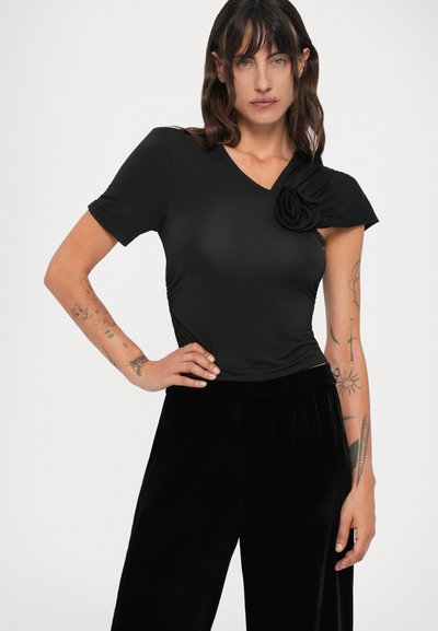 Žena s kestenjastom kosom do ramena, obučena u crni asimetrični top s detaljima od tkanine u obliku cvijeta i crne baršunaste hlače, pozira s rukom na boku.