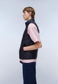 Mladá osoba otočená doľava, oblečená v čiernej prešívanej veste cez svetloružové tričko s krátkym rukávom, s jednou rukou v vrecku vesty.