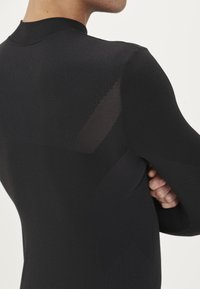 Svart sporttop i ett texturerat tyg, med genomskinliga paneler på ryggen och längs ärmarna. Den åtsittande designen framhäver form och struktur.