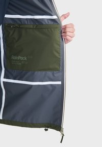 Veste imperméable en matériau gris, avec une poche intérieure verte, des rayures blanches et des détails de fermeture éclair en bas.