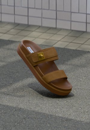 Sandale mule en daim marron avec deux brides, une boucle dorée et une semelle en caoutchouc striée sur l'asphalte près d'un trottoir pavé.