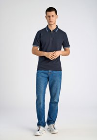 Marinblå polotröja med vita och blå ränder på kragen, kombinerad med blå jeans och vita sneakers. Modellen står upprätt med händerna knäppta.