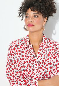 Camicia button-up con base bianca, caratterizzata da una stampa di cuori rossi, dettagli neri e maniche lunghe. Tessuto morbido con una consistenza liscia.