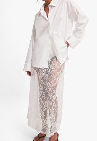 Mujer vestida con una camisa blanca de botones de gran tamaño y una larga falda de encaje blanca transparente con un patrón floral, de pie con una mano en el bolsillo.