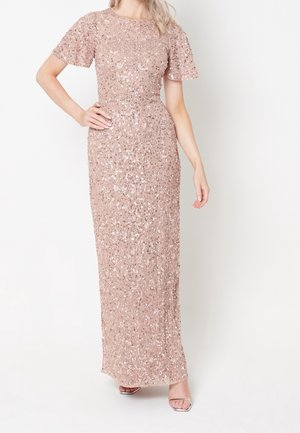 Frau trägt ein bodenlanges, kurzärmeliges, rosafarbenes Pailletten-Kleid mit Blumenmustern und silbernen offenen Schuhen, posiert mit einer Hand in der Hüfte.