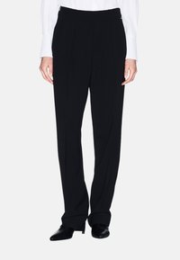 Zwarte broek van een glatte stof, met een hoge taille, plooien en zijzakken. Gepaard met puntige zwarte schoenen.