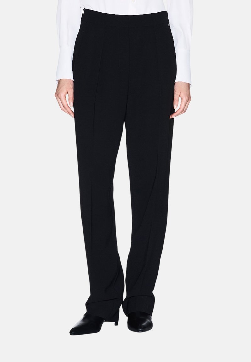 Zwarte broek van een glatte stof, met een hoge taille, plooien en zijzakken. Gepaard met puntige zwarte schoenen.