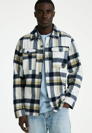 Mann trägt ein weiß-navy-gelb kariertes Hemd über einem hellblauen T-Shirt, kombiniert mit hellblauen Jeans, steht mit einer Hand in der Hosentasche.